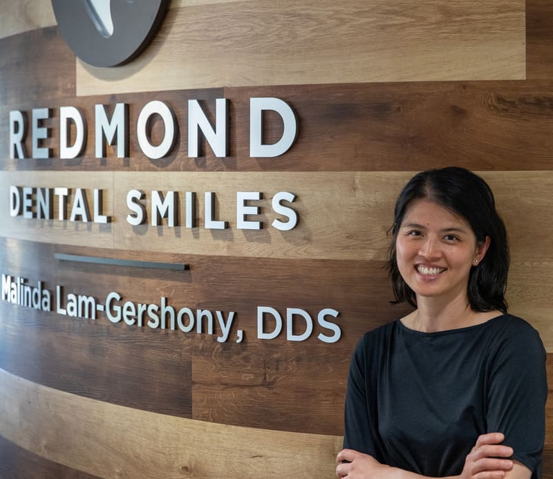 Dr. Malinda Lam-Gershony standing in front of the Redmond Dental Smiles logo wall