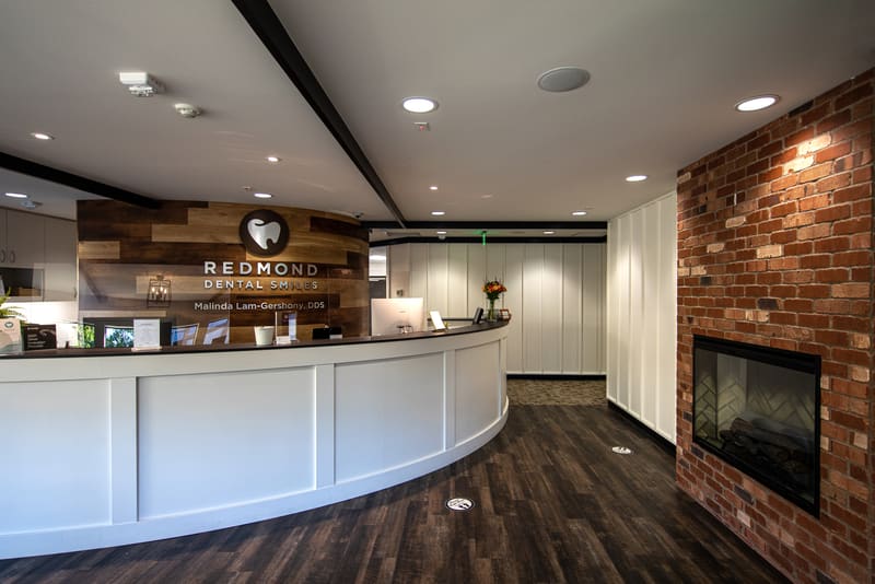 Reception desk at Redmond Dental Smiles with the practice logo on a reclaimed-wood wall and a brick fireplace